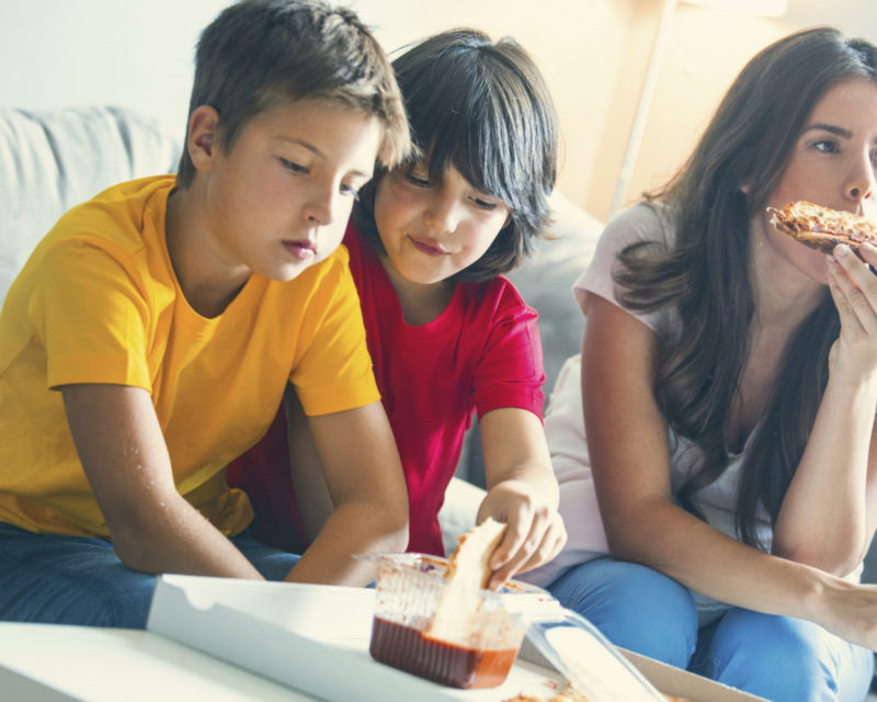 Closeup of two elementary aged boys having a bite with their mother. They are in the living and watching tv while eating. Boys are wearing red and yellow t-shirts. Toned image.